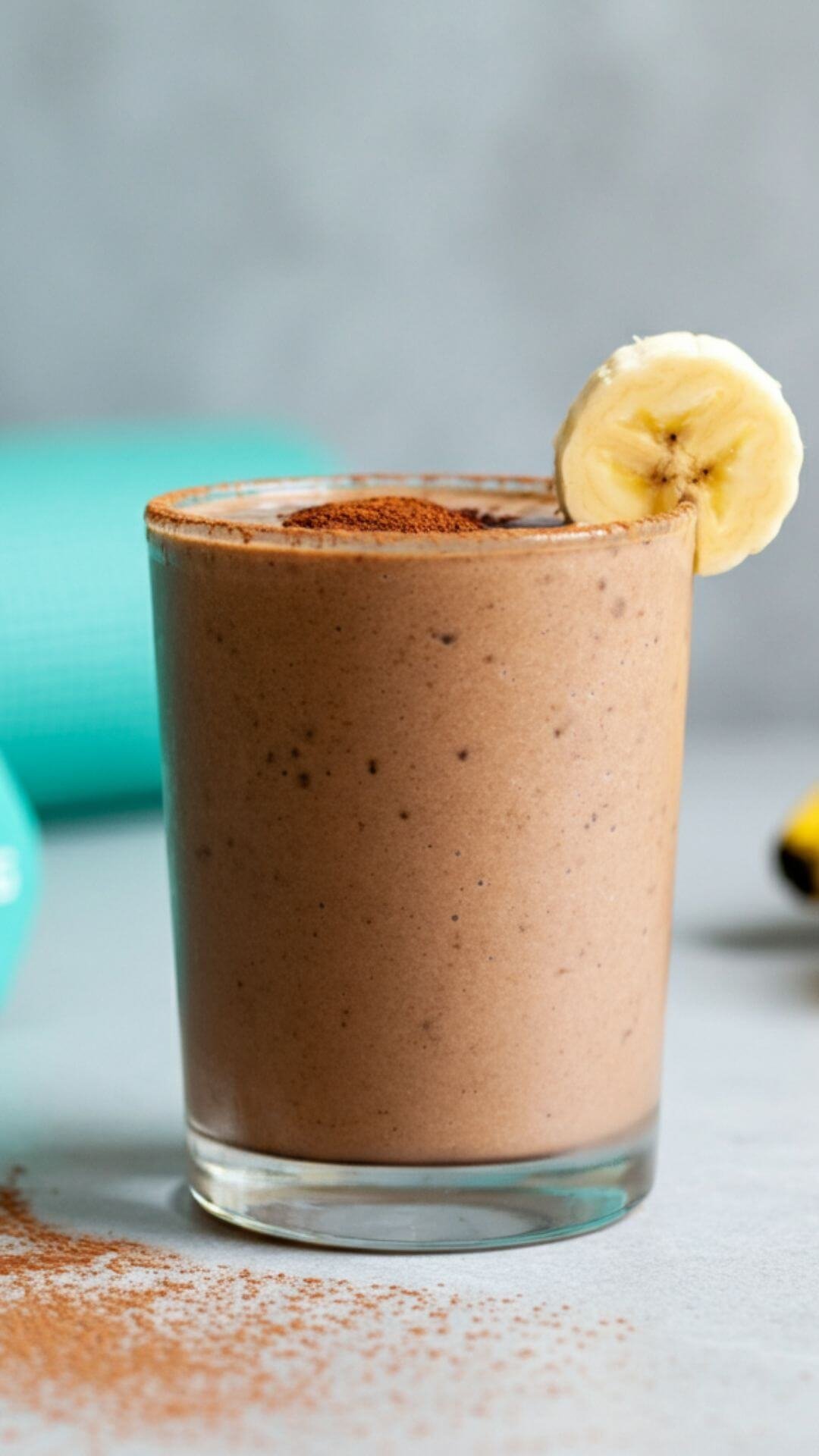 A vibrant and appetizing image of a chocolate banana protein smoothie served in a clear glass, topped with a sprinkle of cocoa powder and a small banana slice on the rim.The background should be clean and bright, with a hint of fitness or health-focused elements like a yoga mat or dumbbells subtly placed.