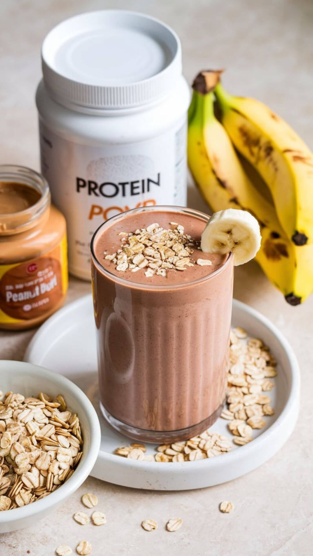 A tall glass of chocolate-colored smoothie sits in the center, garnished with a sprinkle of oats and a banana slice on the rim. Surrounding the glass are the ingredients: a jar of peanut butter, a container of protein powder, a bunch of bananas, a bowl of oats. All placed on the white dish.