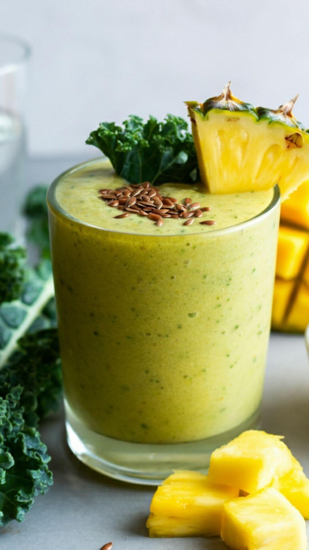 A vibrant tropical smoothie in a clear glass, garnished with a small kale leaf and pineapple chunk. Surround the glass with fresh kale leaves, pineapple chunks, mango slices, and a small bowl of flaxseeds. The background is bright and clean, with a hint of coconut water in a nearby glass or bottle for a tropical vibe.