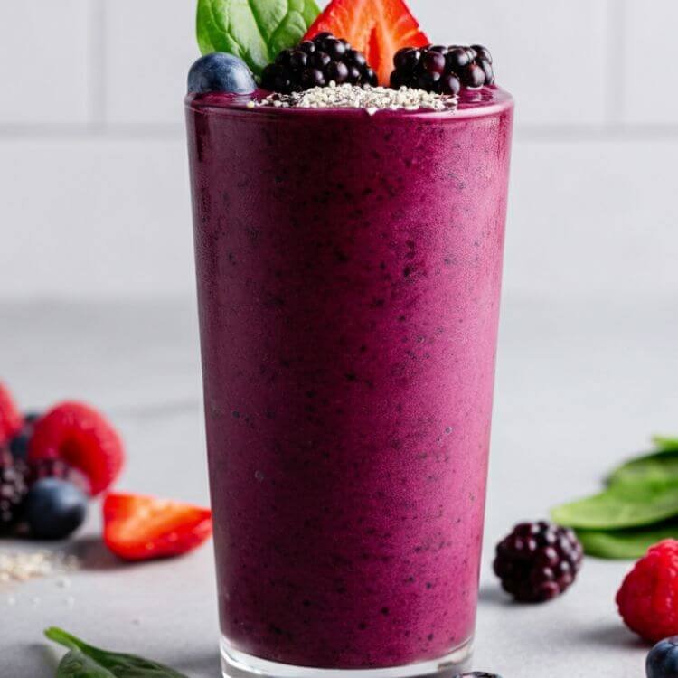 A vibrant smoothie in a clear tall glass, rich purple in color with a smooth texture, garnished with fresh mixed berries, a sprinkle of acai powder, and a small spinach leaf.