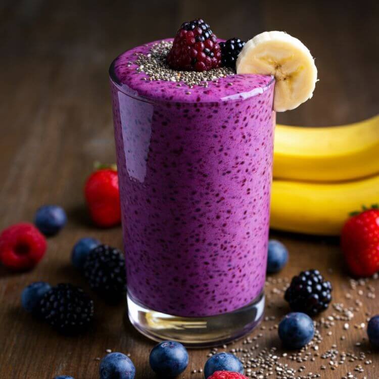 A vibrant smoothie placed in a clear tall glass, showcasing a rich purple hue from mixed berries. The glass is garnished with a sprinkle of chia seeds on top and a small slice of banana on the rim.