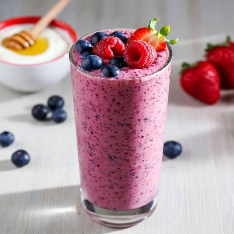 A vibrant and refreshing berry smoothie in a clear tall glass, garnished with fresh blueberries, strawberries, and raspberries on top. The smoothie has a rich, deep pink-purple color, with a creamy texture visible through the glass.