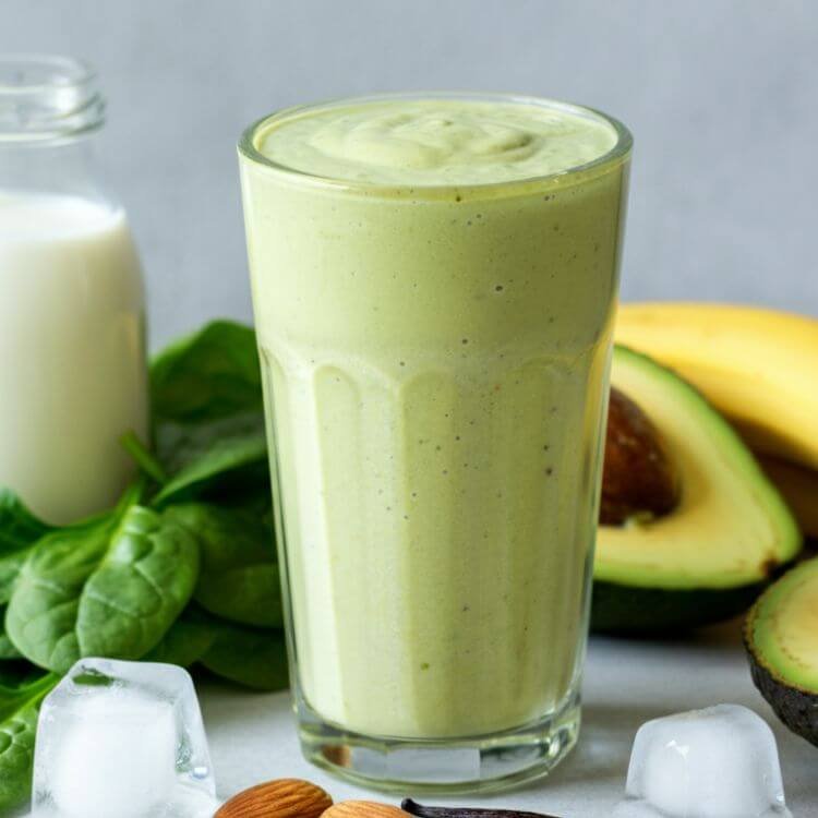 A vibrant smoothie shot showcasing a creamy light green drink in a clear tall glass with a smooth texture. Surround the glass with fresh ingredients: half an avocado, a handful of bright green spinach leaves, half a banana, a small bottle of almond milk, and a sprinkle of vanilla beans. Add a few ice cubes nearby for a refreshing vibe. Use soft natural lighting to enhance the fresh and healthy feel.