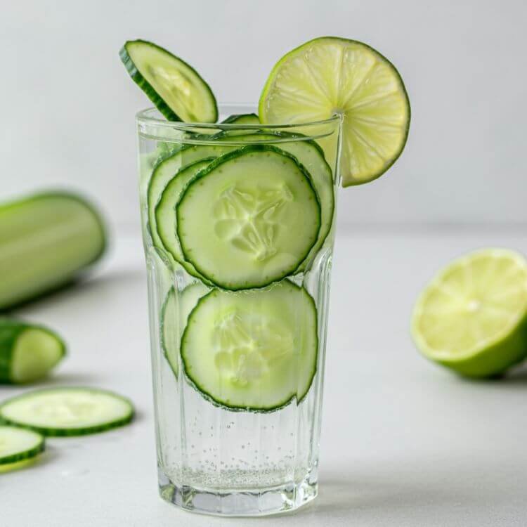 A refreshing glass of cucumber lime soda, featuring thin cucumber slices floating in clear soda water, with a lime wedge on the rim of the glass