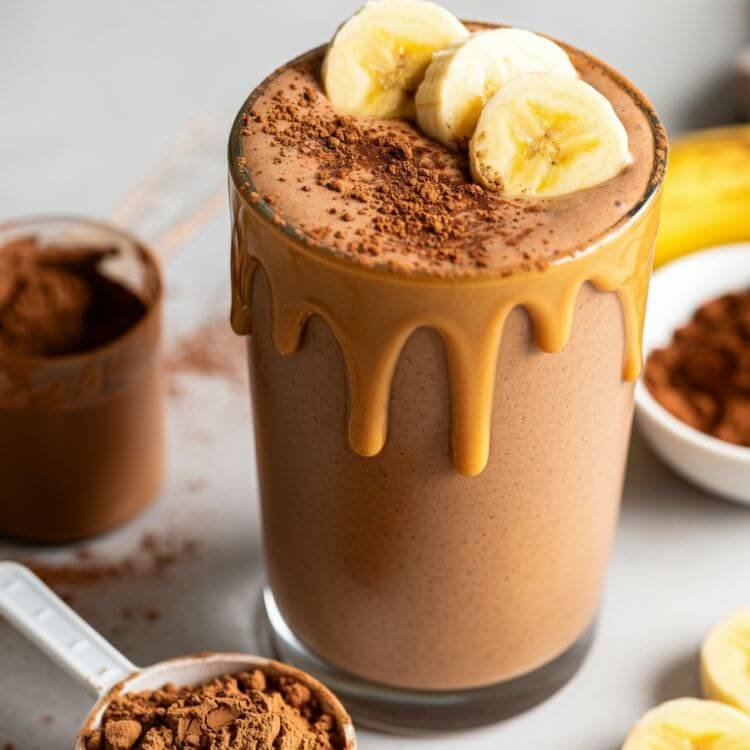 A vibrant close -up of a smoothie setup: a tall glass of rich, creamy chocolate-peanut butter smoothie topped with a drizzle of peanut butter, a sprinkle of cocoa powder, and banana slices, all plaaced on white dish. Surround it with the ingredients—chocolate protein powder in a scoop, a banana, and a small dish of cocoa powder. Warm, natural lighting enhances the rich, indulgent tones.