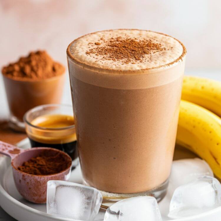 A vibrant close-up of a glass of creamy, frothy mocha protein shake topped with a sprinkle of cocoa powder, surrounded by a scoop of protein powder, a shot of espresso, a halved frozen banana, and a few ice cubes, all placed on white dish. The background is bright and inviting, with soft natural lighting emphasizing the refreshing and energizing vibe of the drink.