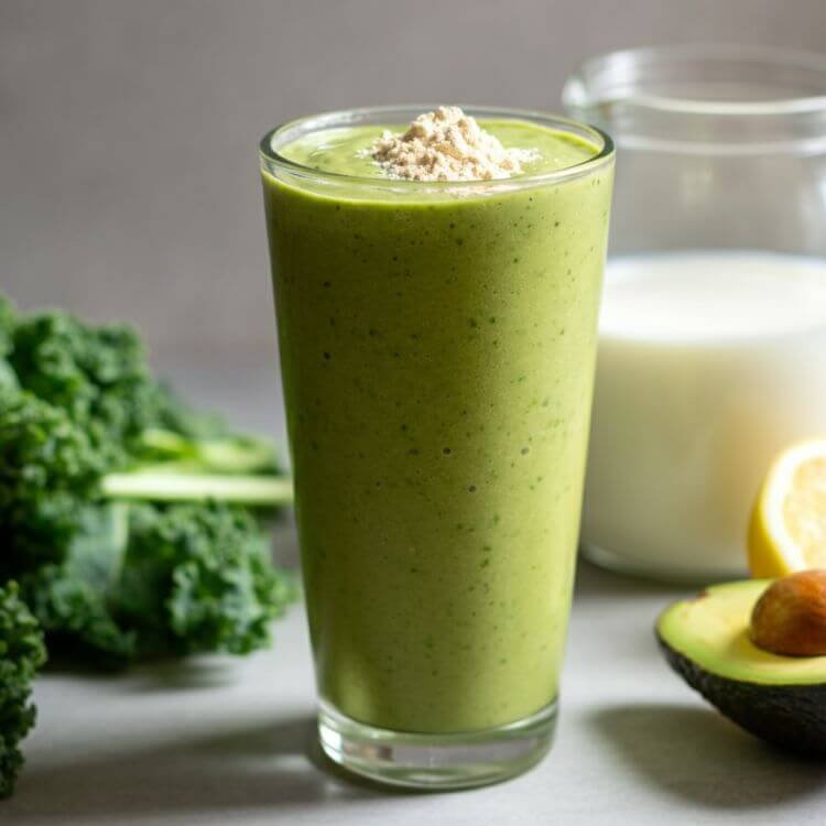 A vibrant smoothie in a clear tall glass, showcasing a creamy green blend. Surround the glass with fresh ingredients: a scoop of vanilla protein powder, leafy kale or spinach, half an avocado, a cup of almond milk, and a sliced lemon. The setting is bright and clean, emphasizing health and freshness.